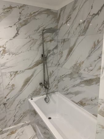 Een prachtig ontworpen luxe badkamer, met een witte strakke badkuip, versierd met marmeren tegels. De elegantie van het marmer wordt versterkt door licht, die een moderne en uitnodigende sfeer aan de ruimte toevoegen. Gedomineerd door tinten grijs en wit, brengt de afbeelding een gevoel van verfijning en moderniteit over. De badkamer bevat een luxueus bad. De verlichting speelt een cruciale rol bij het accentueren van de complexe details van het marmer en het algehele ontwerp van de badkamer. De nauwgezette indeling van de ruimte suggereert een mix van esthetische aantrekkingskracht met praktisch gebruik, waardoor het niet alleen een kamer maar een toevluchtsoord is. De aandacht voor detail in de keuze van materialen, weerspiegelt een doordacht ontwerp dat is bedoeld om een serene en weelderige badkamerervaring te creëren. Tegelwerk door Dobruy Bouwbedrijf, partner van SV Stukadoors