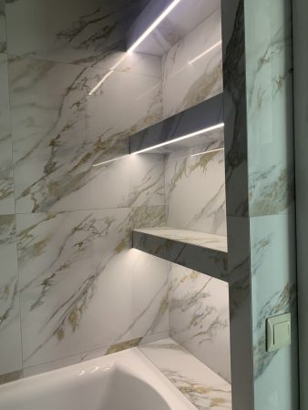 Een prachtig ontworpen badkamer, met prominent een douchehoek versierd met marmeren tegels. De elegantie van het marmer wordt versterkt door zorgvuldig gepositioneerde lampen, die een warme en uitnodigende sfeer aan de ruimte toevoegen. Gedomineerd door tinten grijs en groen, brengt de afbeelding een gevoel van verfijning en moderniteit over. De badkamer bevat een bad, wat het luxueuze gevoel van de kamer benadrukt. De aanwezigheid van sanitaire voorzieningen duidt op functionaliteit naast stijl. De verlichting, zowel ingebed in het plafond als geplaatst op een plank, speelt een cruciale rol bij het accentueren van de complexe details van het marmer en het algehele ontwerp van de badkamer. De nauwgezette indeling van de ruimte suggereert een mix van esthetische aantrekkingskracht met praktisch gebruik, waardoor het niet alleen een kamer maar een toevluchtsoord is. De aandacht voor detail, van de keuze van materialen tot de plaatsing van lampen, weerspiegelt een doordacht ontwerp dat is bedoeld om een serene en weelderige badkamerervaring te creëren. Tegelwerk door Dobruy Bouwbedrijf